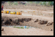 Water jugs lined up collecting water for a borehole near a filthy river bottom